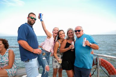 Smiling group of adults cheering with canned drinks on a sunny boat deck over coastal waters, wearing summer clothes and sunglasses