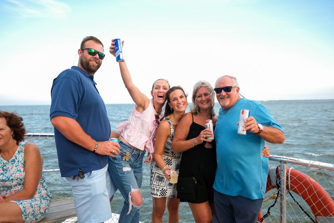Smiling group of adults cheering with canned drinks on a sunny boat deck over coastal waters, wearing summer clothes and sunglasses