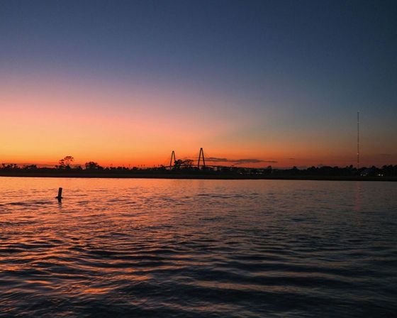 Serene orange-purple sunset over rippled water, silhouetted bridge and distant radio tower reflecting across a calm river estuary.