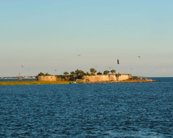 Historic coastal fort on a palm-topped island in a calm blue bay at golden hour, flag flying, seabirds overhead and a small boat by the shore.