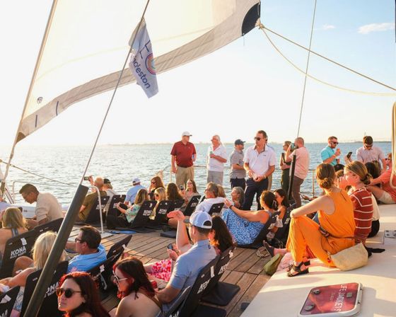 Group enjoying a sunset sail on a wooden-decked sailboat, lounging in deck chairs and on the deck under white sails with calm ocean and horizon