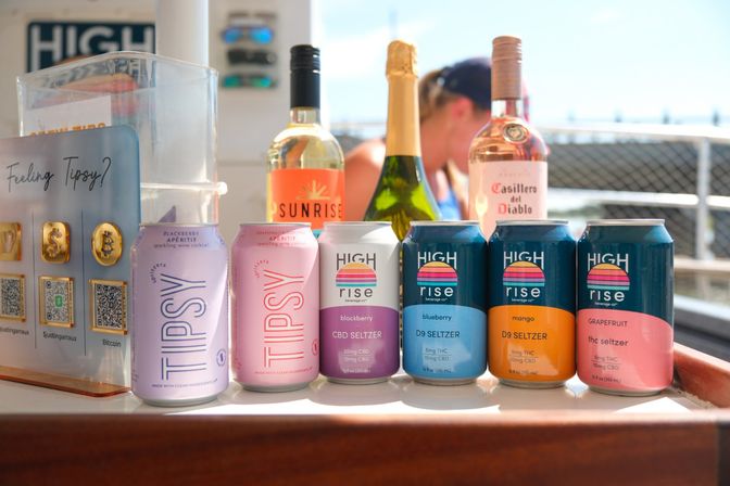 Sun-soaked row of colorful CBD/THC seltzer cans in berry, mango and grapefruit flavors with wine bottles behind on a waterfront bar counter.