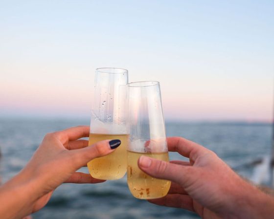 Two hands clinking champagne flutes over the ocean at a pastel sunset, a sparkling-wine toast on the water