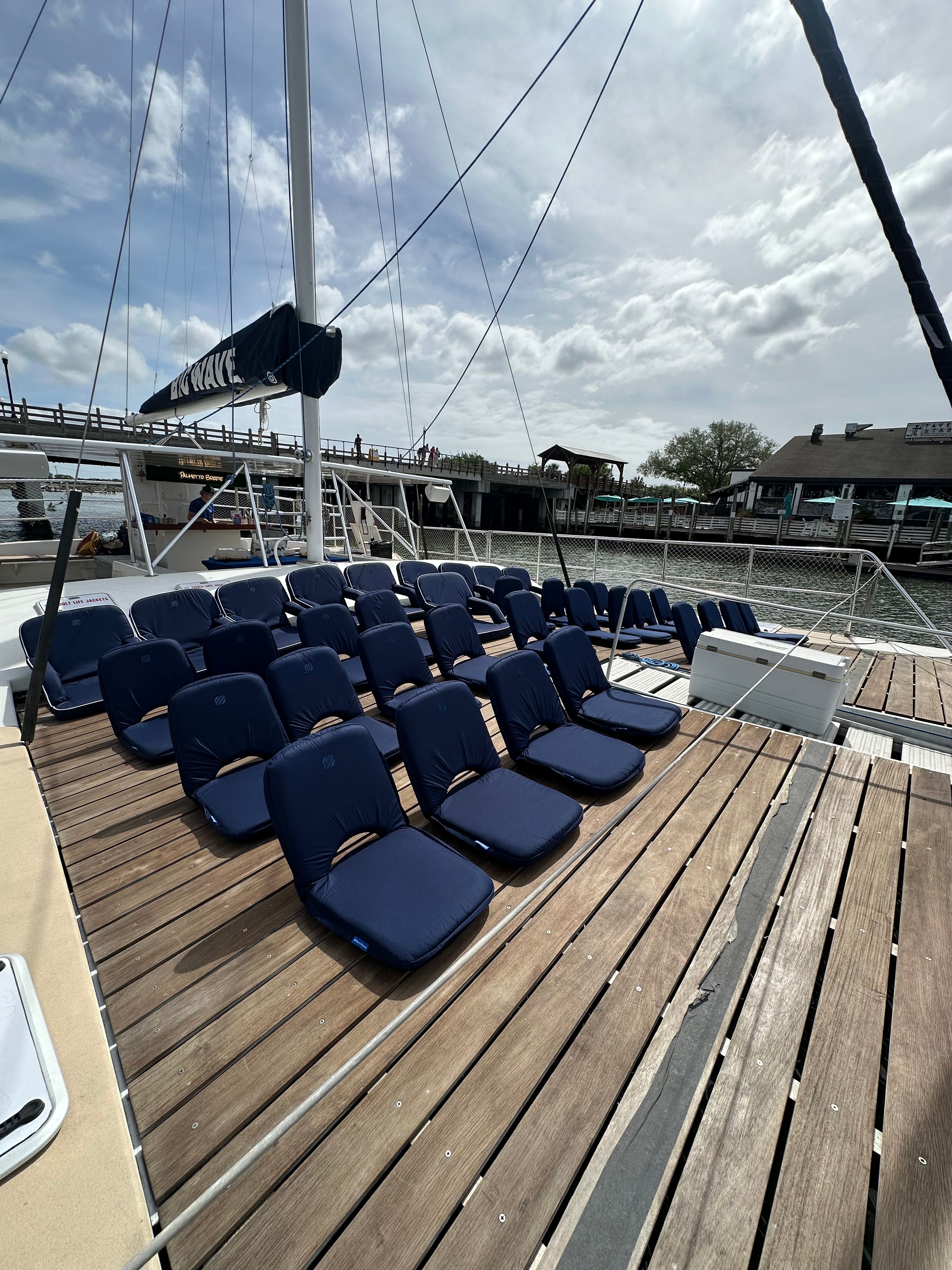Rows of navy blue cushioned seats arranged on a wooden catamaran deck at a marina, with mast and rigging overhead, a pedestrian pier and waterfront buildings under a partly cloudy sky