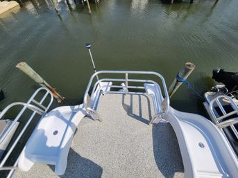 Top-down view of a sunlit boat swim platform with textured deck, stainless-steel railing and boarding ladder, tied to wooden pilings in a calm marina harbor.