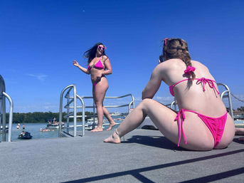 Two women in pink bikinis on a lakeside dock under a bright blue sky — one posing in heart-shaped sunglasses while the other photographs her, with boats and swimmers in the background on a sunny summer day.