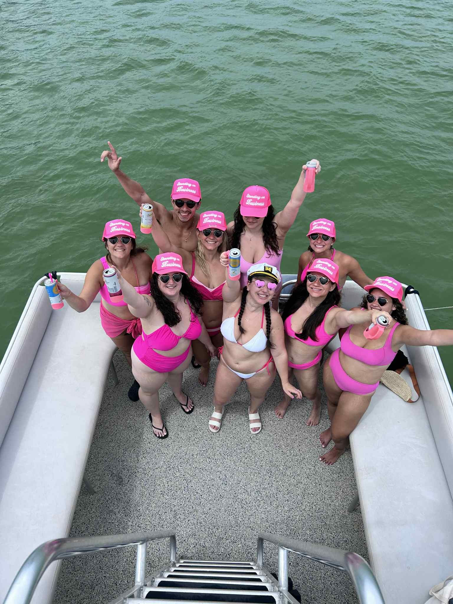 Group of friends in matching pink swimsuits and caps cheering with drinks on a pontoon boat, smiling up at the camera over green water.