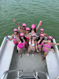 Group of friends in matching pink swimsuits and caps cheering with drinks on a pontoon boat, smiling up at the camera over green water.