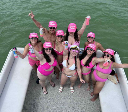 Group of friends in matching pink swimsuits and caps cheering with drinks on a pontoon boat, smiling up at the camera over green water.