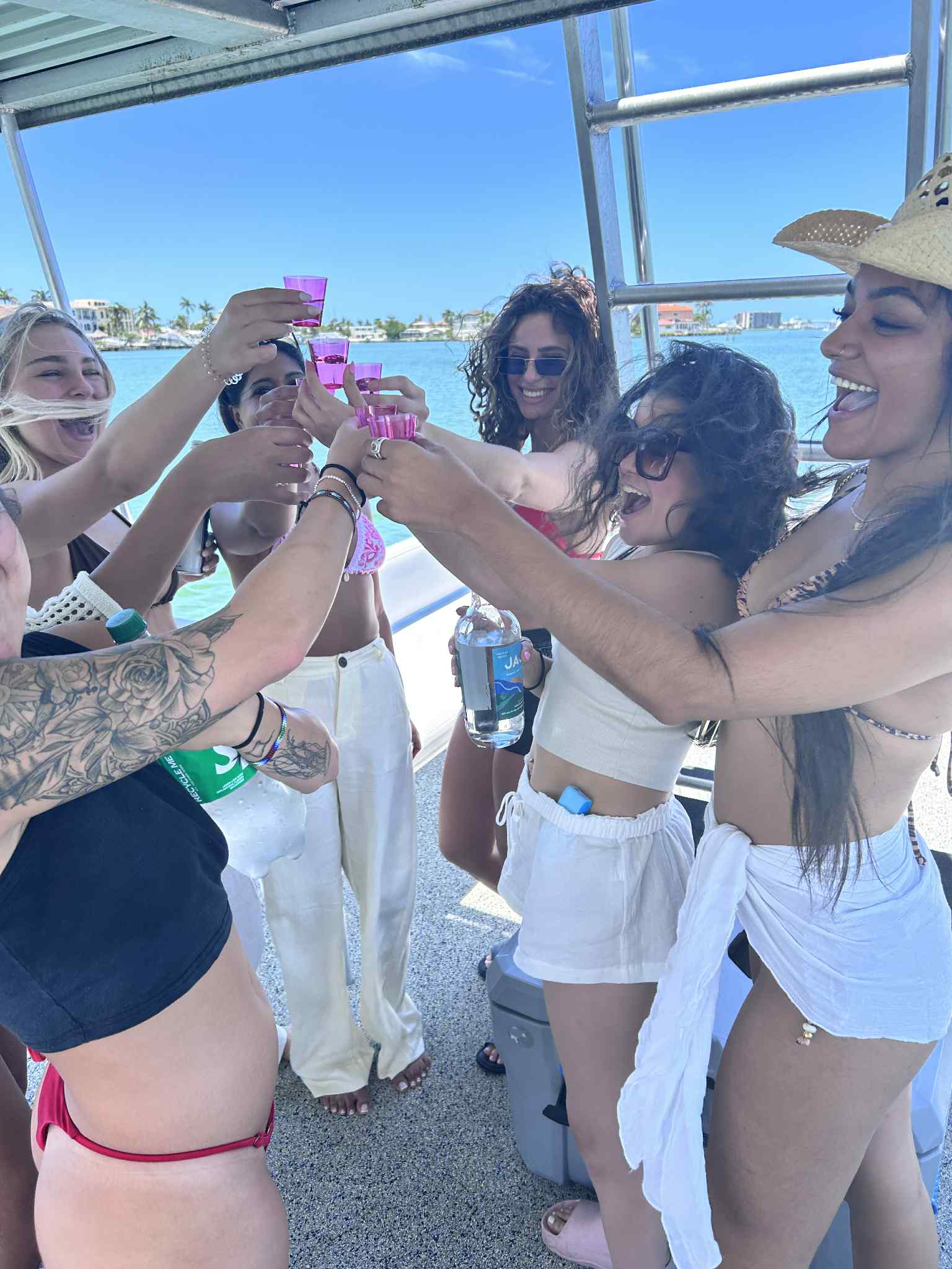 Group of friends on a sunny boat party toasting with pink shot glasses over clear turquoise water and a palm-lined coastal skyline — vacation vibes, summer boat celebration.