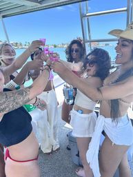 Group of friends on a sunny boat party toasting with pink shot glasses over clear turquoise water and a palm-lined coastal skyline — vacation vibes, summer boat celebration.