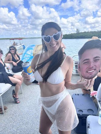 Smiling woman in a white bikini and heart-shaped sunglasses holding a canned drink and a large face cutout on a pontoon boat with friends, blue sky and fluffy clouds over a calm lake