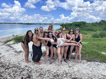 Group of friends in swimsuits posing on a white shell-covered coastal beach by calm blue ocean, lush green dunes and puffy cloud sky