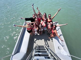 Group of women in red swimsuits and sunglasses cheering and holding drinks aboard a pontoon boat on green lake water, photo taken from the ladder above.