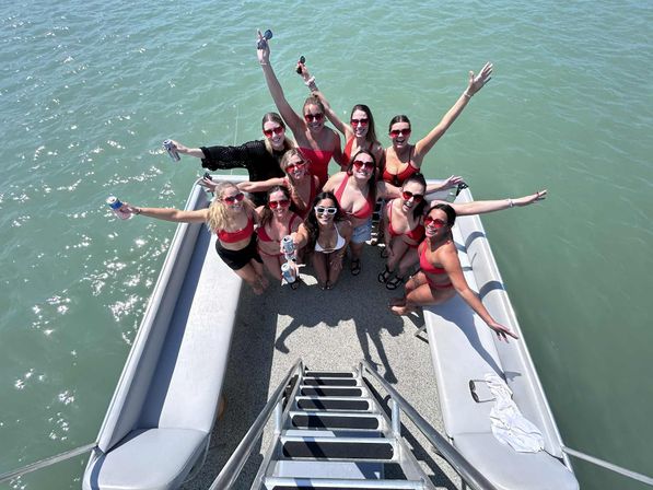 Group of women in red swimsuits and sunglasses cheering and holding drinks aboard a pontoon boat on green lake water, photo taken from the ladder above.