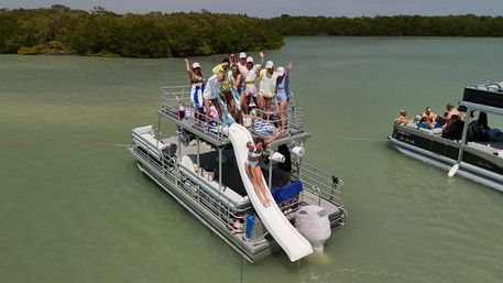 Two-level pontoon boat with a white waterslide as a woman in a white bikini slides down and a cheering group in swimwear and matching caps gathers on the upper deck, anchored in shallow green water near a mangrove-lined shoreline.