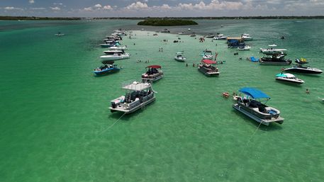 Drone view of boats anchored around a shallow turquoise sandbar with swimmers enjoying a sunny coastal day