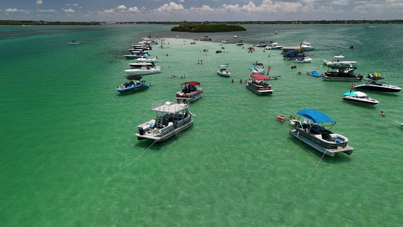Drone view of boats anchored around a shallow turquoise sandbar with swimmers enjoying a sunny coastal day