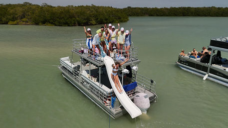 Pontoon party boat with an upper-deck waterslide as a woman slides into shallow green coastal lagoon, friends waving on deck near mangrove shoreline and a nearby pontoon.