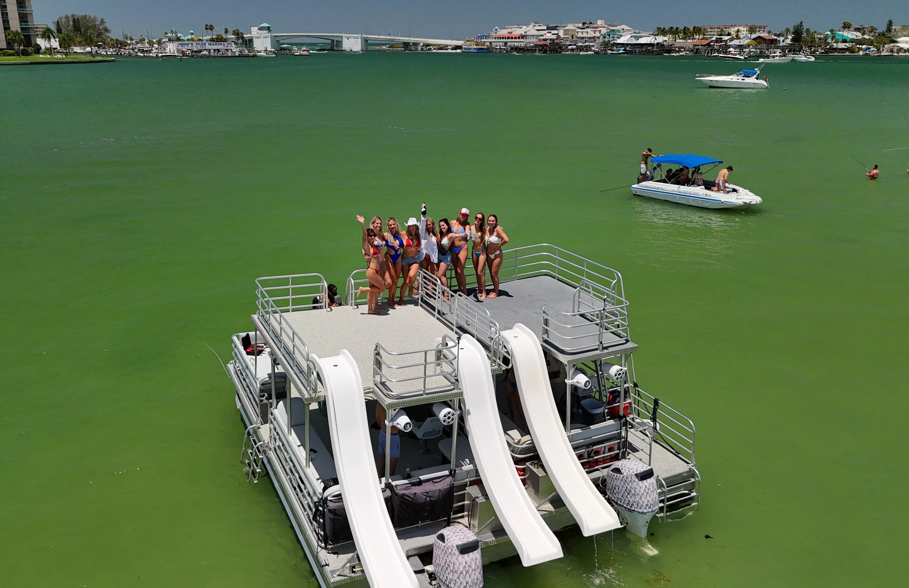 Cheering group on a double-deck pontoon party boat with three white water slides in a turquoise bay, small boats nearby and a coastal town on the horizon on a sunny summer day