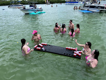 Friends in swimsuits playing beer pong on a floating table in shallow green bay water, surrounded by anchored boats and other partygoers