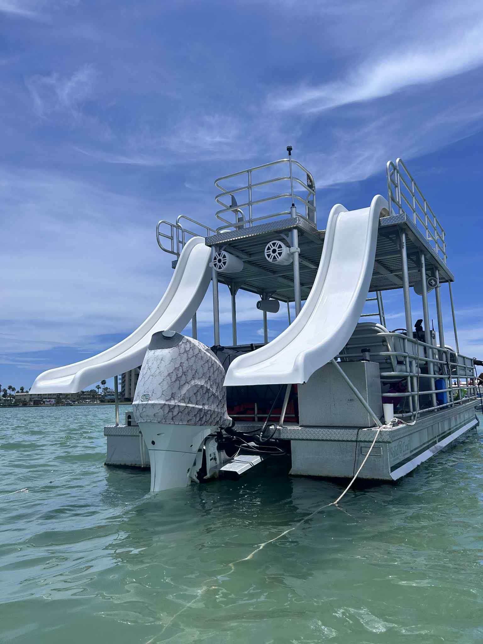 Playful two-level pontoon boat with twin white water slides and outboard motor bobbing in clear turquoise coastal water under a bright blue sky