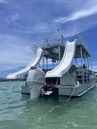 Playful two-level pontoon boat with twin white water slides and outboard motor bobbing in clear turquoise coastal water under a bright blue sky
