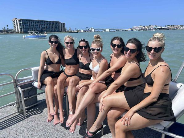 Seven women in bikinis and sunglasses smiling together on a boat in a sunny coastal bay with waterfront buildings and a bridge in the background.