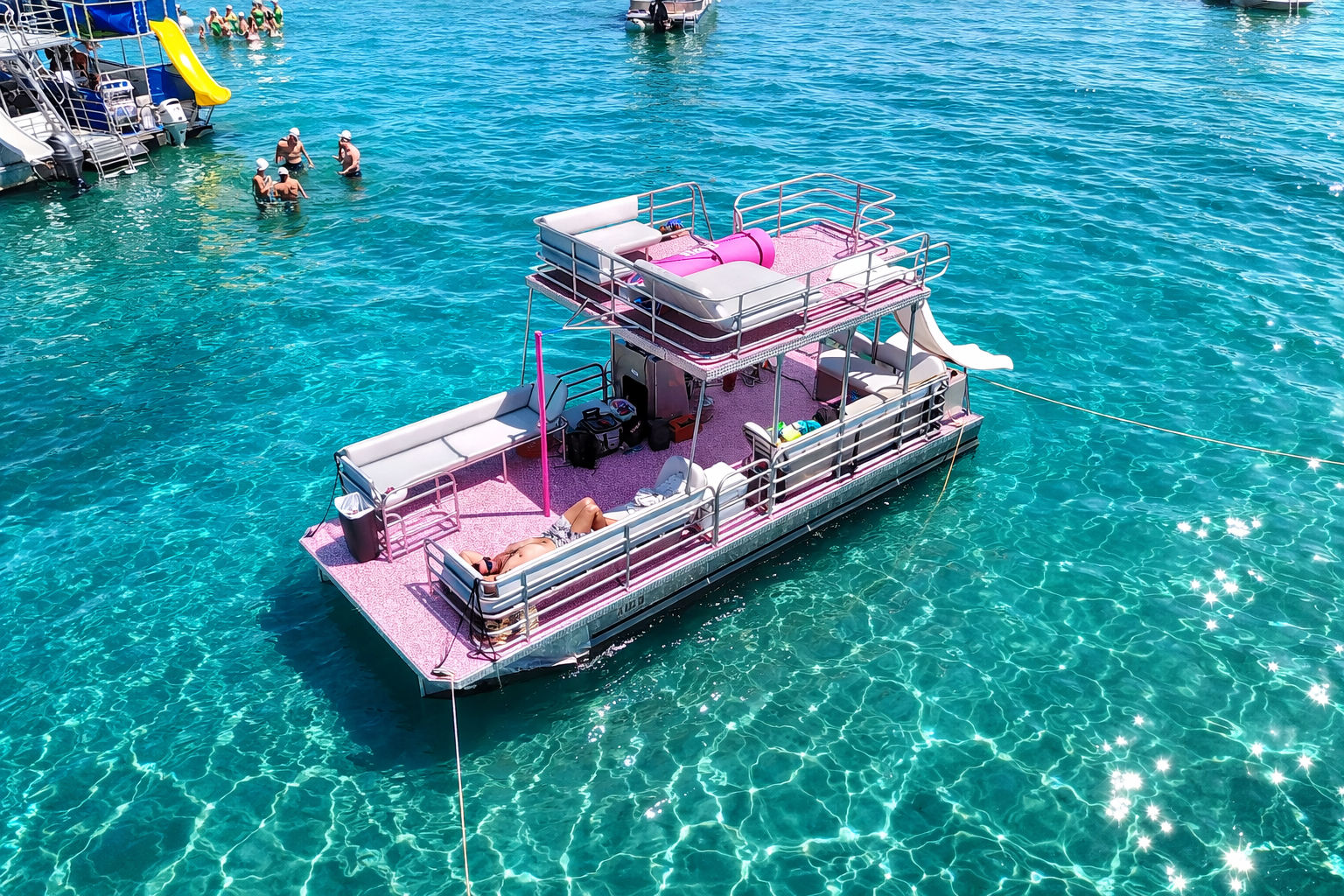 Pink double-decker pontoon boat with lounge seating and a slide anchored in crystal-clear turquoise water, sunbathers on deck and swimmers nearby on a bright summer day.