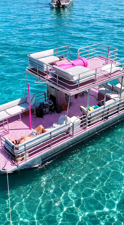 Pink double-decker pontoon boat with lounge seating and a slide anchored in crystal-clear turquoise water, sunbathers on deck and swimmers nearby on a bright summer day.
