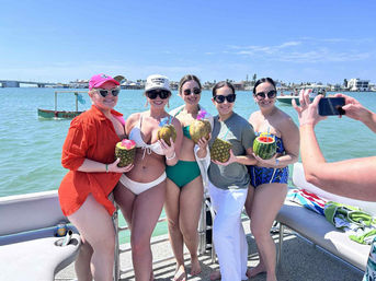 Five women in swimsuits on a sunny boat in turquoise coastal waters, smiling and holding tropical fruit cocktails served in coconuts, pineapples and a hollowed watermelon.