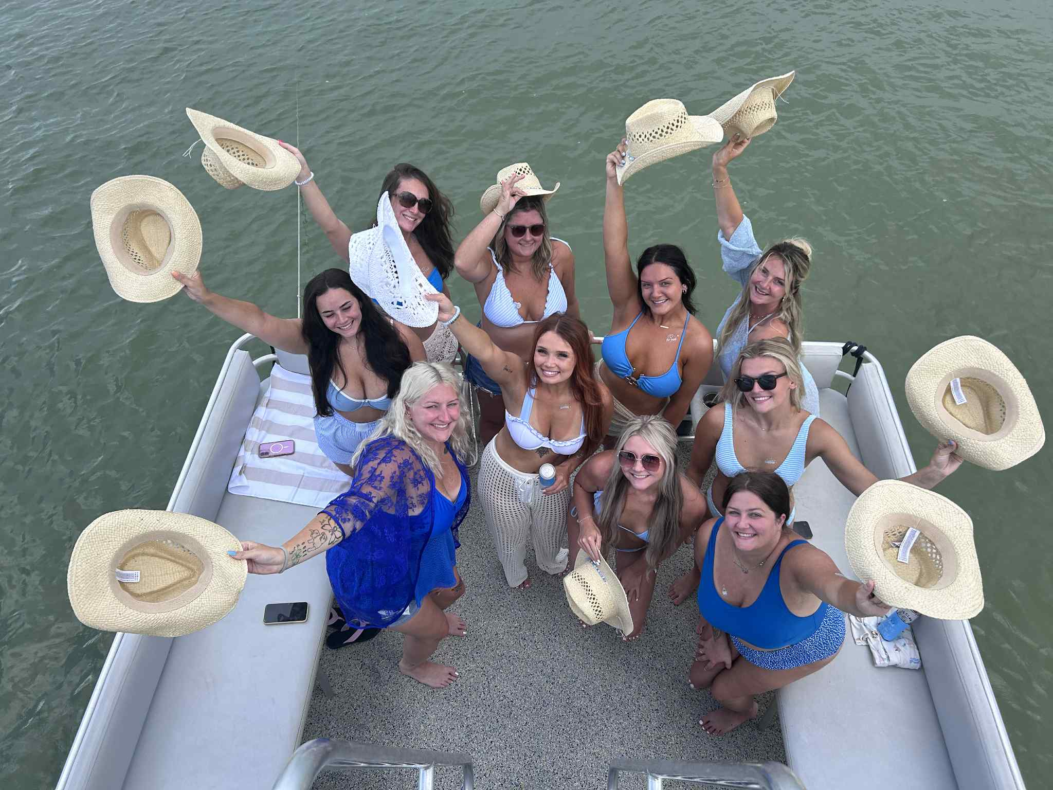 Group of women in bikinis and swimsuits on a pontoon boat on a lake, smiling and waving straw cowboy hats overhead for a sunny summer boating party.