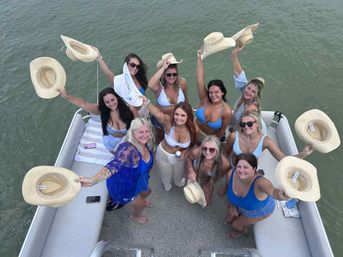 Group of women in bikinis and swimsuits on a pontoon boat on a lake, smiling and waving straw cowboy hats overhead for a sunny summer boating party.