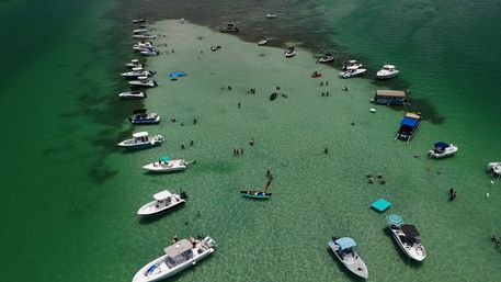 Aerial view of boats anchored around a shallow sandbar in crystal-clear turquoise water, with people wading, swimming, and lounging on floats.