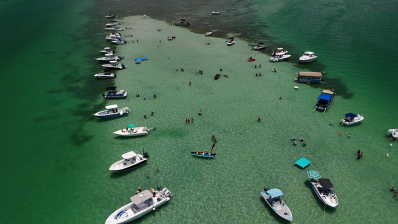 Aerial view of boats anchored around a shallow sandbar in crystal-clear turquoise water, with people wading, swimming, and lounging on floats.