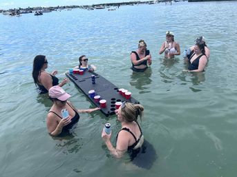 Group of women in swimsuits playing beer-pong on a floating table in shallow coastal bay water, holding canned drinks with boats and shoreline visible in the background