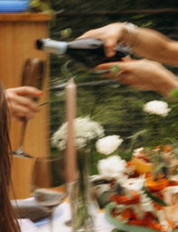Motion-blurred hands popping a champagne bottle at an outdoor garden table set with a pink taper candle, white and orange floral centerpiece, and a guest holding a wine glass.