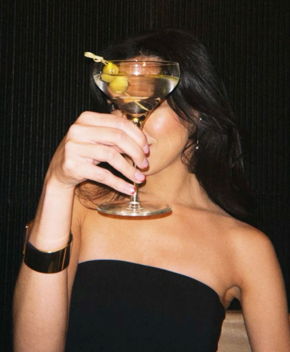 Person in a strapless black dress and gold cuff holding a martini glass with two olives up to cover their face against a dark backdrop — chic cocktail, night-out vibe in a cocktail lounge.
