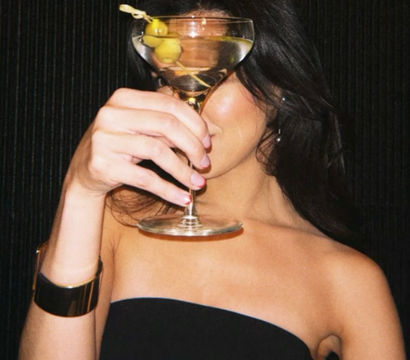 Person in a strapless black dress and gold cuff holding a martini glass with two olives up to cover their face against a dark backdrop — chic cocktail, night-out vibe in a cocktail lounge.
