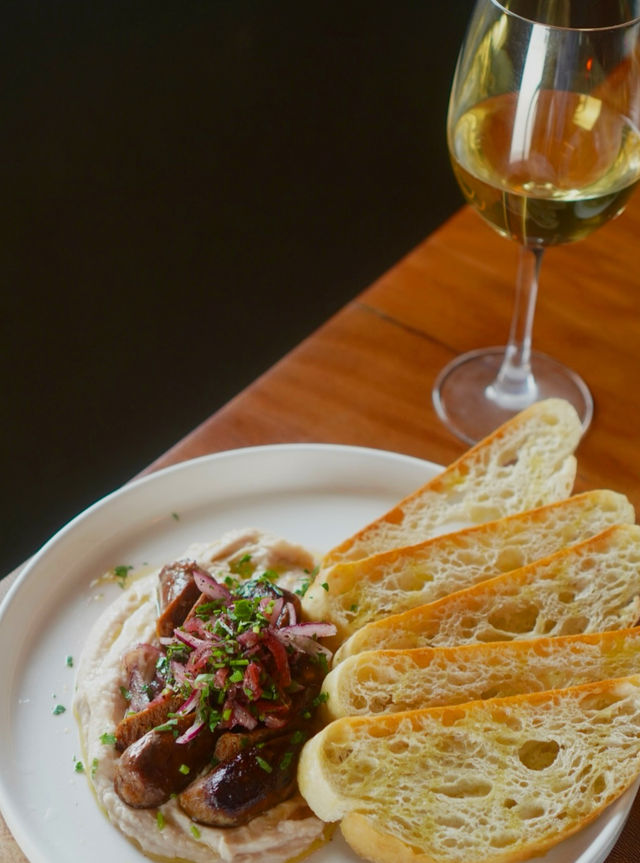Plate of grilled sausages on creamy white bean purée topped with pickled red onions, served with toasted baguette slices and a glass of white wine on a wooden bistro table