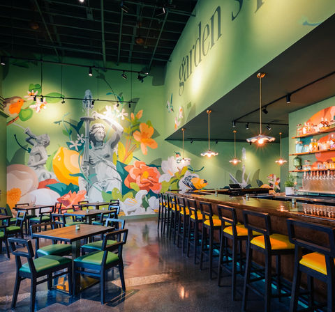 Garden-themed cafe interior with a large colorful floral mural and classical statue motif, green walls, wooden bar with yellow-cushioned stools, pendant lights and wooden dining tables on a polished concrete floor.