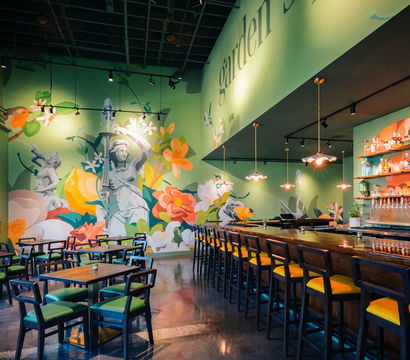 Garden-themed cafe interior with a large colorful floral mural and classical statue motif, green walls, wooden bar with yellow-cushioned stools, pendant lights and wooden dining tables on a polished concrete floor.