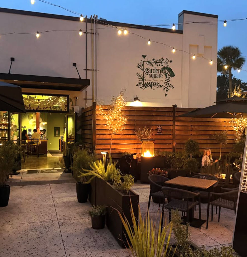 Cozy evening outdoor patio dining with string lights, potted plants, wooden privacy fence and a glowing fire pit by the restaurant entrance.