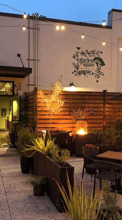 Cozy evening outdoor patio dining with string lights, potted plants, wooden privacy fence and a glowing fire pit by the restaurant entrance.