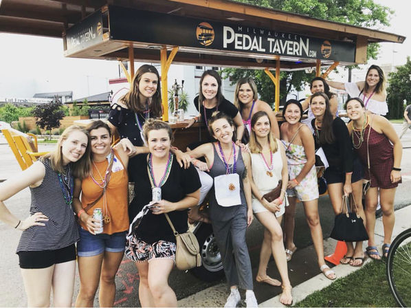 Group of smiling women wearing colorful bead necklaces posing by a pedal-powered party bar on a sunny Milwaukee street, festive outdoor celebration vibe.