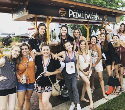 Group of smiling women wearing colorful bead necklaces posing by a pedal-powered party bar on a sunny Milwaukee street, festive outdoor celebration vibe.