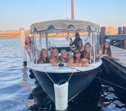 Smiling group of women enjoying a summer boat outing docked at an urban waterfront harbor on a covered leisure boat with a small American flag.