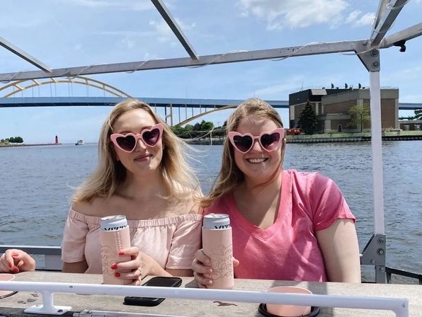 Two friends in pink outfits and heart-shaped sunglasses smiling and holding canned drinks on a sunny boat ride with an arched bridge and lighthouse over a river