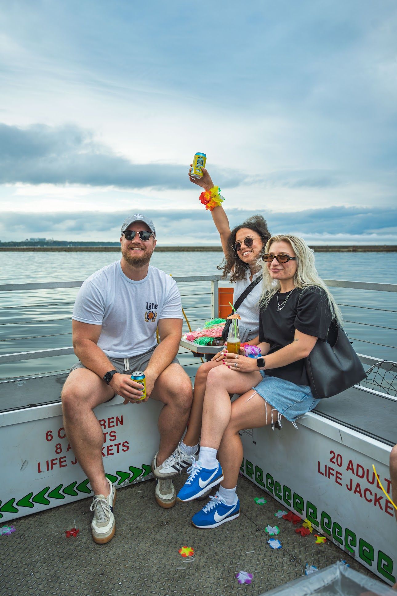 Three friends enjoying a small boat party on calm waterfront waters, holding drinks and wearing colorful leis under an overcast summer sky.