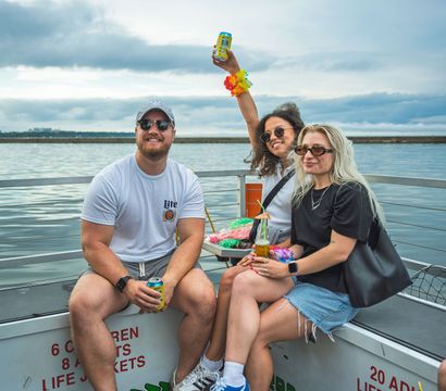 Three friends enjoying a small boat party on calm waterfront waters, holding drinks and wearing colorful leis under an overcast summer sky.